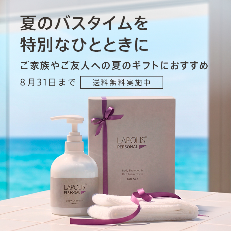 夏のギフトセットが海辺のテーブルに置かれた写真。ボディシャンプーと箱入りギフト、紫のリボン付き。なお、背景には海が広がる夏の雰囲気。